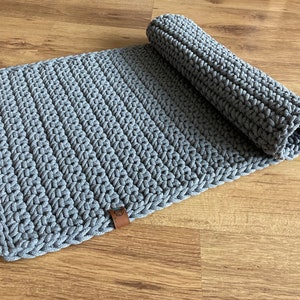 Pode incluir: Um tapete de crochê cinza com uma etiqueta de couro marrom. O tapete está enrolado e está em um chão de madeira.