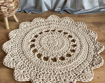May include: A beige crocheted round rug with a scalloped edge. The rug has a circular pattern in the centre.