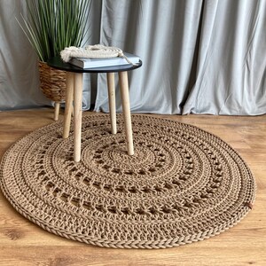 Può includere: Un tappeto rotondo in crochet beige con un motivo circolare. Un piccolo tavolo nero con gambe in legno è posizionato sopra il tappeto.