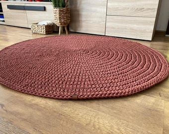 Könnte beinhalten: Ein runder, handgehäkelter Teppich in einem gedämpften, rötlich-braunen Farbton. Die Textur des Teppichs ist sichtbar, mit konzentrischen Kreisen, die ein strukturiertes Muster bilden. Der Teppich liegt auf einem Holzboden.