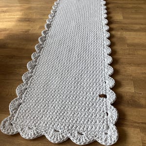 May include: A rectangular, light gray crochet rug with a scalloped edge. The rug is made of thick yarn and has a textured surface. The rug is approximately 40 inches long and 20 inches wide.