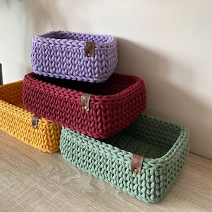 Puede incluir: Cuatro cestas rectangulares de ganchillo en tonos amarillo, burdeos, verde y morado están apiladas. Cada cesta tiene una etiqueta de cuero. Las cestas están hechas de hilo grueso y son adecuadas para guardar o decorar.