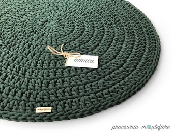May include: A dark green, circular, crocheted rug. The rug is made of thick yarn and has a textured surface. A small tag with the word "omnia" is attached with twine. A small metal tag with the brand name is also visible.