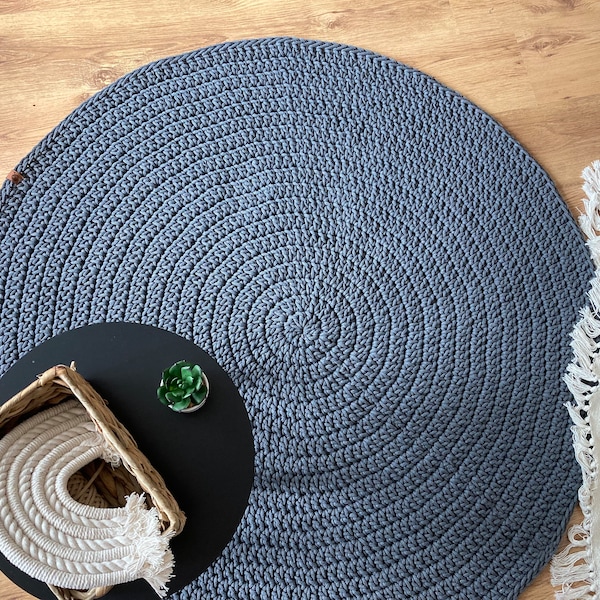 May include: A large, round, crocheted rug in a solid gray color. The rug has a spiral design and is placed on a wooden floor. A small black table with a basket and a plant is in the foreground.