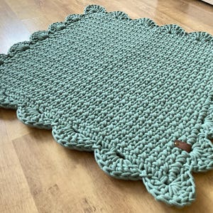May include: A sage green, rectangular crochet rug with a scalloped edge. The handmade rug is made of thick yarn and features a small, brown tag. The rug is displayed on a wooden surface.