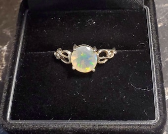 Puede incluir: Un anillo de plata con una gran piedra preciosa de ópalo redonda. El ópalo muestra un juego de colores con destellos verdes y amarillos. El anillo está engastado en una banda de plata decorativa, presentado en una caja negra.