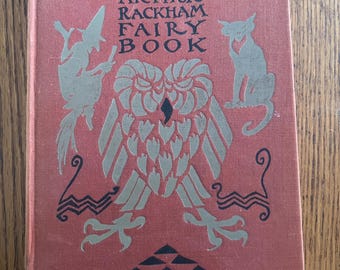 Puede incluir: Un libro antiguo titulado "The Arthur Rackham Fairy Book" con una cubierta roja. La portada presenta ilustraciones plateadas de un búho, una bruja y un gato. Los elementos decorativos incluyen formas geométricas y líneas estilizadas.