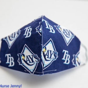 May include: A navy blue face mask with a repeating pattern of the Tampa Bay Rays logo in white and light blue. The mask has white elastic ear loops and is made of fabric. The text "TB RAYS" is visible on the mask.