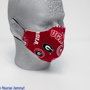 May include: A red and white face mask featuring the University of Georgia Bulldogs logo and text. The mask is made of fabric and has black elastic ear loops. The mask is displayed on a mannequin head.