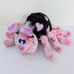 May include: A plush toy spider with a pink body, legs, and ears. The spider's back is black with pink heart-shaped designs. The toy has blue eyes and a pink mouth.