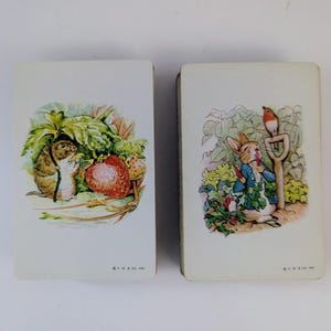 May include: Two rectangular playing cards featuring classic illustrations. The left card shows a brown mouse near a large red strawberry. The right card depicts a rabbit in a blue jacket eating a carrot with a small bird perched on a garden tool.