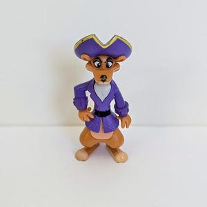 May include: A small, plastic figurine of a cartoon character. The figure is a brown animal wearing a purple pirate-style hat with gold trim and a matching jacket. The figure has a white shirt, black belt, and brown pants.