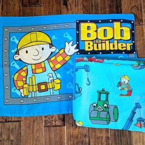 May include: A light blue Bob the Builder themed bedding set. The set includes a pillowcase with Bob the Builder and a sheet with construction vehicles. The Bob the Builder logo is also visible.