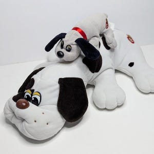 May include: A large white plush dog with black spots and dark brown ears, featuring a smaller grey plush puppy with black ears and a red collar resting on its back. Both plush toys have embroidered facial features and small red tags attached.