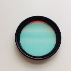 May include: A black camera lens filter with a light blue and red coloured glass insert.