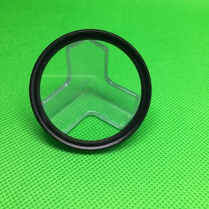 May include: A black camera lens filter with a clear plastic insert. The filter is sitting on a green surface.