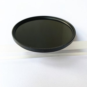 May include: A circular, black camera lens filter with a dark, reflective glass surface. The filter has a thin, textured black metal rim. The item is set against a white background, with a clear, rectangular object partially visible.