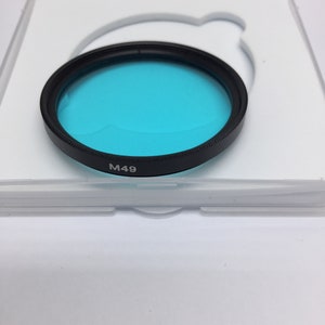 Könnte beinhalten: Ein blauer Filter für ein Kameraobjektiv in einem weißen Kunststoffgehäuse. Der Filter ist mit "M49" gekennzeichnet.