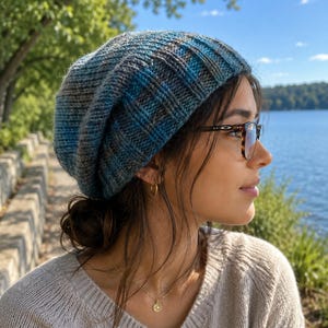 Puede incluir: Un gorro de punto azul y gris con un diseño acanalado. El gorro está hecho de hilo suave y tiene un ajuste holgado. El gorro lo lleva una persona con gafas y un suéter de color claro.
