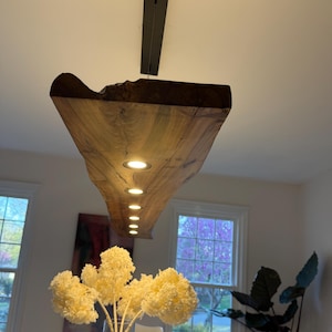 May include: A wooden, live-edge light fixture with recessed lights hangs above a dining table. The fixture is made of dark brown wood. A vase of yellow flowers sits on the table. The room has white walls and windows.