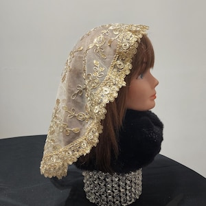 May include: A gold-colored lace veil with floral embroidery and a scalloped edge. The veil is draped over a mannequin head with brown hair and a black fur collar. The base is a decorative, crystal-studded cylinder.