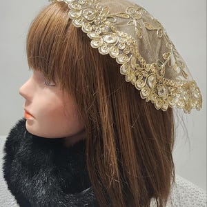 May include: A sheer, gold-colored lace head covering with a scalloped edge. The lace is delicate and intricate, with a floral pattern.