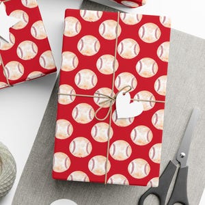 Puede incluir: Varias cajas de regalo envueltas en papel rojo con un patrón de béisbol. Cada regalo está atado con hilo y adornado con una etiqueta de papel blanco. También se ven una madeja de hilo y unas tijeras.