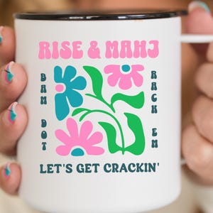 May include: White enamel mug with a black rim and handle. The mug features the text "RISE & MHJ" in pink, and "LET'S GET CRACKIN'" in navy blue. A floral design with blue and pink flowers and green leaves is also present.
