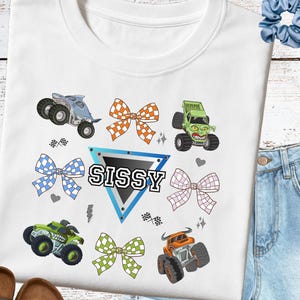 May include: White sweatshirt featuring colorful monster truck graphics and checkered bows. The word "SISSY" is displayed in a blue triangle. Additional graphics include hearts, stars, and checkered flags.