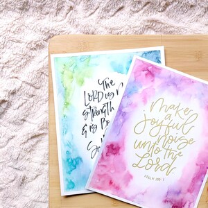 May include: Two watercolor prints with inspirational bible verses. The first print has a blue and green watercolor background with the text "The Lord is my strength and is my song". The second print has a pink and purple watercolor background with the text "Make a joyful noise unto the Lord. Psalm 100:1".