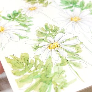 Pode incluir: Ilustra&ccedil;&atilde;o em aquarela de margaridas brancas com centros amarelos e folhas verdes. As flores s&atilde;o retratadas com linhas delicadas e suaves aguadas de aquarela, criando uma est&eacute;tica fresca e natural. A obra de arte est&aacute; em um fundo branco.