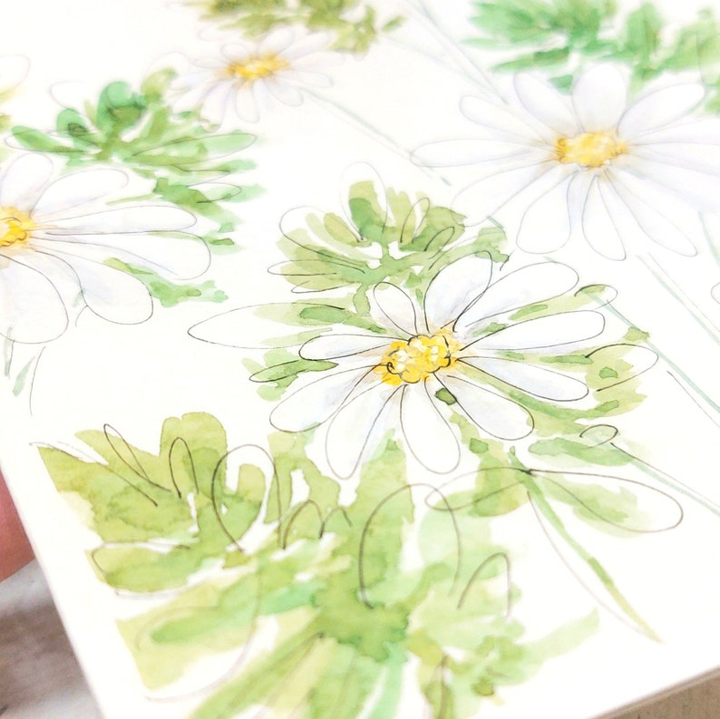 Pode incluir: Ilustra&ccedil;&atilde;o em aquarela de margaridas brancas com centros amarelos e folhas verdes. As flores s&atilde;o retratadas com linhas delicadas e suaves aguadas de aquarela, criando uma est&eacute;tica fresca e natural. A obra de arte est&aacute; em um fundo branco.