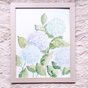 Pode incluir: Pintura em aquarela de hort&ecirc;nsias em uma moldura de madeira clara. A obra apresenta hort&ecirc;nsias azuis, roxas e verdes com pinceladas delicadas. As folhas s&atilde;o verde-suaves e o fundo &eacute; branco.