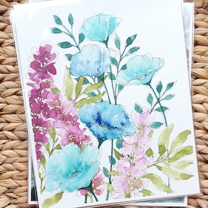 Pode incluir: Impress&atilde;o de arte floral em aquarela com papoulas azuis e turquesas, flores rosa e roxas e folhas verdes. A obra de arte est&aacute; em papel branco e &eacute; protegida por uma capa de pl&aacute;stico transparente. A impress&atilde;o &eacute; exibida em um fundo de vime tecido.