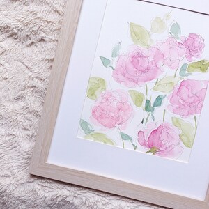 Pode incluir: Pintura em aquarela de rosas cor-de-rosa e folhas verdes em uma moldura branca e uma moldura de madeira clara. A obra floral apresenta pinceladas delicadas e uma est&eacute;tica suave e arejada, ideal para decora&ccedil;&atilde;o de interiores.