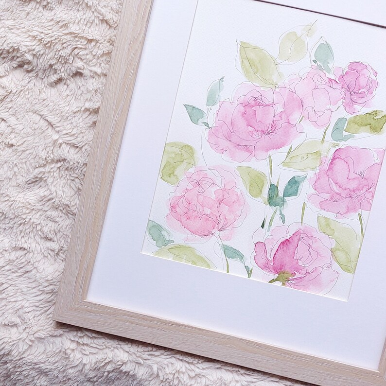 Pode incluir: Pintura em aquarela de rosas cor-de-rosa e folhas verdes em uma moldura branca e uma moldura de madeira clara. A obra floral apresenta pinceladas delicadas e uma est&eacute;tica suave e arejada, ideal para decora&ccedil;&atilde;o de interiores.