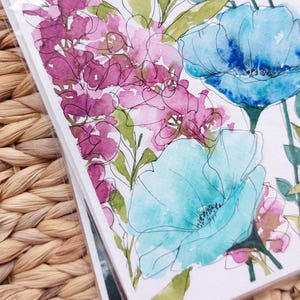 Pode incluir: Impress&atilde;o de arte floral em aquarela com flores azuis e roxas e folhas verdes. A obra de arte tem um estilo delicado, pintado &agrave; m&atilde;o, com detalhes finos em linha preta. As flores est&atilde;o em um fundo branco.