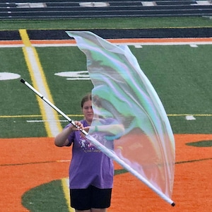 Peut inclure: Une personne tient un drapeau avec un mât argenté et un drapeau translucide et irisé. La personne porte un t-shirt violet et un short noir, debout sur un terrain vert avec des marquages orange.