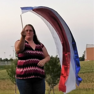 Puede incluir: Una persona sostiene una bandera de guardia de color con rayas rojas, blancas y azules. La bandera está curvada y ondeando. La persona lleva una camiseta con estampado rosa y negro y vaqueros azules.