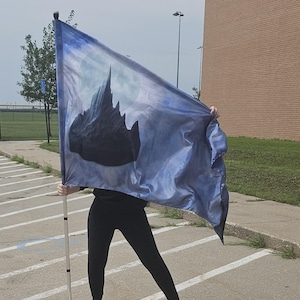 May include: A blue and silver flag with a mountain silhouette design, held by a person. The flag is on a silver pole. The flag's design includes a full moon. The person is wearing black leggings and gray sandals.