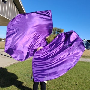 May include: Two purple satin dance flags, also known as worship flags, are held by a person in a grassy field. The flags are flowing in the wind.