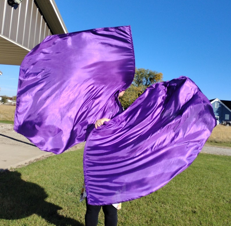May include: Two purple satin dance flags, also known as worship flags, are held by a person in a grassy field. The flags are flowing in the wind.