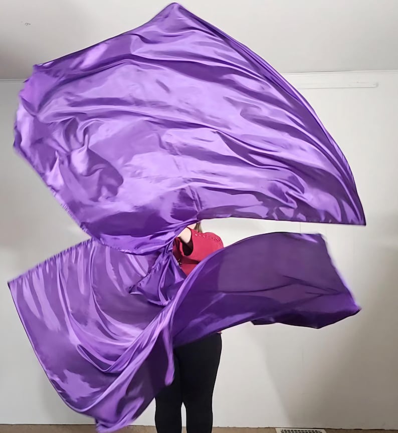 May include: A person is holding two purple dance flags, which are flowing in the air. The flags are made of a silky fabric and are a vibrant purple color.