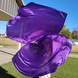 May include: A person holding a large purple fabric flag, flowing in the wind. The flag is made of a shiny, silky material.