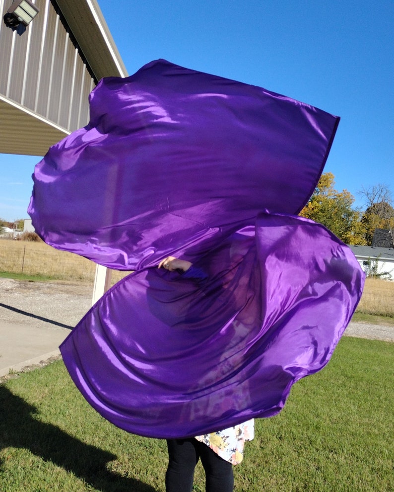 May include: A person holding a large purple fabric flag, flowing in the wind. The flag is made of a shiny, silky material.