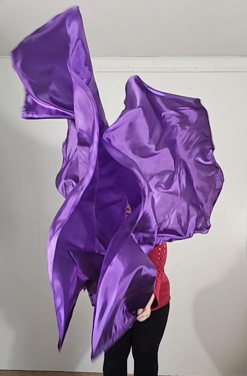 May include: A person is holding and twirling two purple fabric dance flags. The flags are flowing in the air.