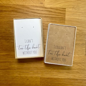 Può includere: Due piccole scatole regalo rettangolari con inserti in cartoncino bianco e marrone. L'inserto bianco recita "I CAN'T tie the knot WITHOUT YOU" in un carattere scritto a mano. L'inserto marrone recita "I COULDN'T tie the knot WITHOUT YOU".