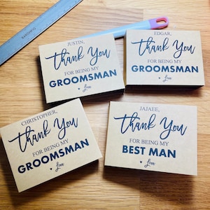 May include: Four brown cards with black text expressing gratitude for being a groomsman or best man. The text on the cards reads "Thank You for being my groomsman" and "Thank You for being my best man". Each card has a small heart with the word "Leon" below it.