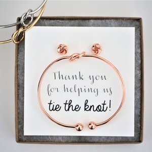 Può includere: Un set di gioielli in oro rosa in una scatola regalo. Il set include un braccialetto annodato, un paio di orecchini a bottone annodati e un biglietto che dice "Grazie per averci aiutato a fare il nodo!"