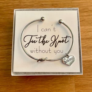 Può includere: Un braccialetto d'argento con un ciondolo a forma di cuore che dice "Bridesmaid". Il braccialetto ha un design a nodo e le parole "Non posso fare il nodo senza di te" sono stampate su una carta bianca dietro il braccialetto.